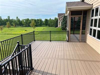 Spacious brown composite deck with black railings, attached to a house with a screened porch, overlooking a wide green lawn and wooded area in the distance in Burnsville.