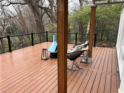 A spacious brown composite deck with black cable railings, attached to a dark gray house. The deck is furnished with modern outdoor chairs and a small blue chiminea, overlooking a wooded backyard in Oak Park Heights.