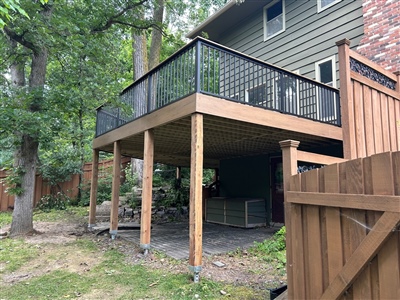 A raised wooden deck supported by tall posts, featuring black railings with mesh panels. The space below the deck is open and shaded, with trees and a wooden fence nearby in Spring Park.