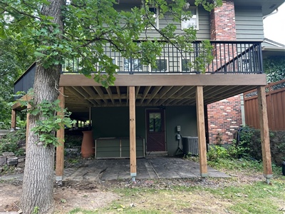 A raised wooden deck supported by several posts, with black railings on the upper level. The space beneath the deck is shaded and partially enclosed, with a door leading into the house and outdoor storage visible in Shakopee.